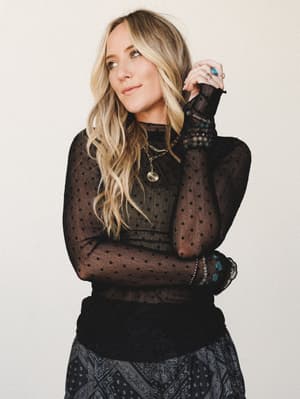 A black sheer polka dot long-sleeve top with ruffled trim is styled with a paisley skirt and layered gold jewelry — the kind of outfit that reads effortlessly cool for a night out without trying too hard.