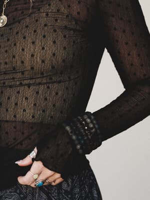 A close-up torso shot of a sheer black polka dot mesh long-sleeve top styled with a paisley skirt, stacked stone bracelets, and layered silver rings — the kind of outfit that feels effortlessly collected and a little mysterious.