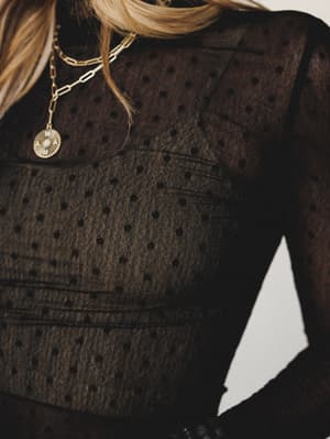 A close-up detail shot of a black polka dot mesh top with delicate seam paneling, styled with layered gold chain necklaces — moody, romantic, and made for a night out.