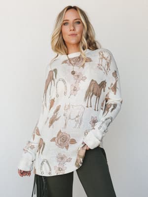 An oversized cream knit sweater printed with a scattered western-inspired motif — horses, roses, horseshoes, and florals in warm taupes and dusty pinks — worn casually over dark pants with a gold pendant necklace and fringe bag, shot against a soft s