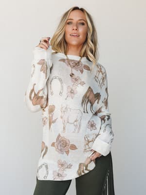An oversized cream knit sweater covered in a scattered western-inspired print — horses, longhorn cattle, horseshoes, and roses — styled with olive leggings and a fringe bag for a lived-in boho-western feel.