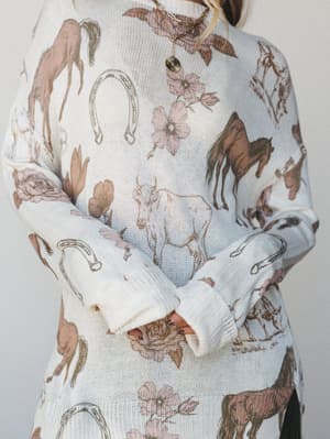 A close-up of a cream knit sweater covered in a vintage-style equestrian print — horses, horseshoes, roses, and florals in soft dusty tones — worn loosely with a delicate gold necklace peeking through.