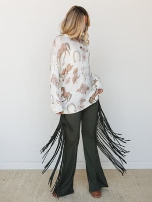 A model shot from behind shows a cream graphic sweater with a western-inspired horse and floral print, paired with dramatic dark olive fringe pants — the kind of outfit that walks into a room and makes a statement without trying.