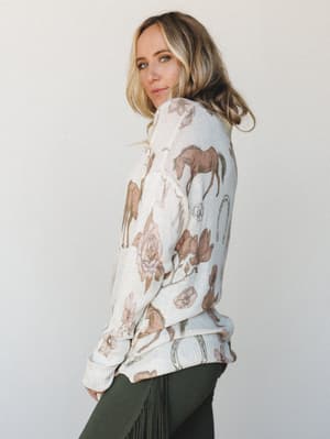 A cream knit sweater with an all-over equestrian-inspired print — horses, horseshoes, and roses in dusty rose and warm brown — worn over olive fringe pants, shot in profile against a soft studio backdrop with a lived-in, story-telling feel.
