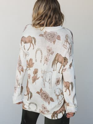 Shot from the back, a model wears an oversized cream knit sweater covered in a whimsical western-inspired print — horses, roses, horseshoes, and florals scattered across the fabric like a mood board for your favorite daydream.