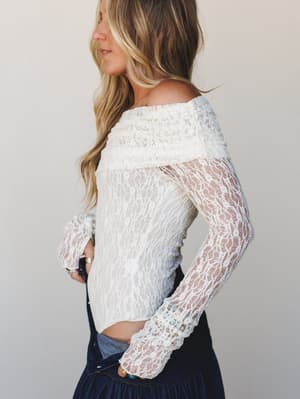 A cream lace bodysuit with an off-shoulder neckline and long sheer sleeves, styled with a dark denim skirt — the kind of piece that works as hard for a date night as it does tucked into jeans for a weekend out.