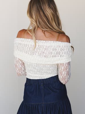 Shot from behind, a model pairs a cream lace off-the-shoulder bodysuit — sheer long sleeves, folded neckline — with a dark denim tiered skirt, the whole look feeling effortlessly romantic against a soft studio backdrop.