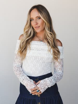 A white lace off-shoulder bodysuit styled with a dark denim tiered skirt, shot against a soft neutral studio backdrop — romantic and a little undone, like she got dressed by instinct and it just worked.