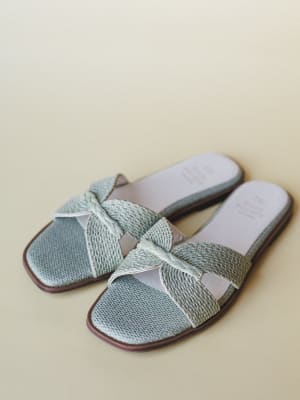 A pair of sage mint woven slide sandals with crossover braided straps and a blush interior, shot against a soft cream background — the kind of sandal that makes you want to book a trip somewhere warm.