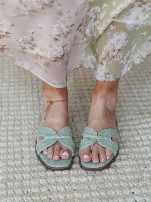 A close-up of sage mint woven slide sandals with a braided knot detail, styled against a jute rug with a flowy floral chiffon skirt grazing the ankles — effortlessly warm-weather and very much the shoes you reach for every single day.