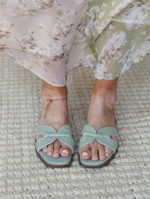 A close-up of sage mint woven slide sandals with a braided knot detail, styled against a jute rug with a flowy floral chiffon skirt grazing the ankles — effortlessly warm-weather and very much the shoes you reach for every single day.