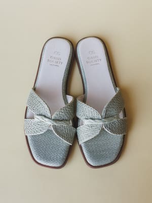 A pair of sage mint woven slide sandals shot from above against a warm cream background — the knotted strap detail and raffia-like texture give them that effortless, grab-and-go summer energy.