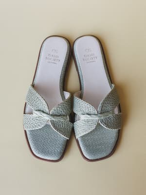 A pair of sage mint woven slide sandals shot from above against a warm cream background — the knotted strap detail and raffia-like texture give them that effortless, grab-and-go summer energy.