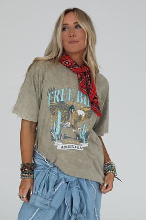 A woman with long blonde hair wears the Fly Free Graphic Tee - Mocha in a boho-chic look, styled with a red bandana, blue denim at her waist, and layered bracelets, standing against a plain white background.