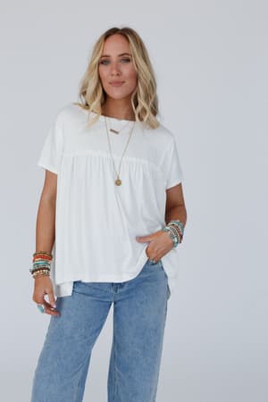 A woman with blonde hair poses confidently against a plain light background, wearing the Sunday Morning Babydoll Top in white with ruffle details, blue jeans, and layered colorful bracelets and necklaces.