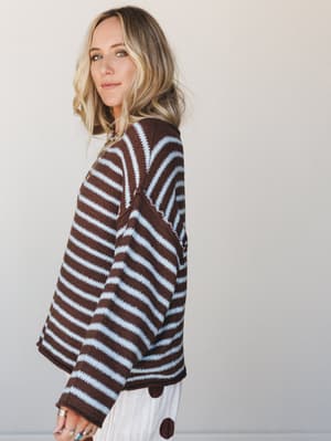 A cozy oversized striped sweater in chocolate and powder blue worn by a blonde model shot from a side angle — the kind of sweater that makes you want to cancel plans and stay somewhere warm.