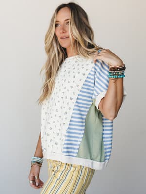 A mixed-print patchwork top in cream with ditsy floral and blue stripe panels styled with yellow stripe pants and layered beaded bracelets — effortlessly eclectic and full of that 'I just threw this on' energy that actually takes real taste.