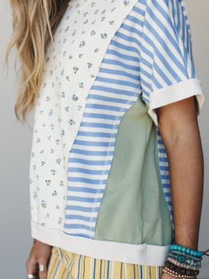 A close-up detail shot of a patchwork top that mixes ditsy floral and blue-and-white stripe panels with a sage color-block side, styled with stacked beaded bracelets and a peek of yellow striped bottoms — the kind of top that looks like it was made f