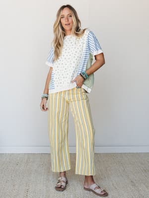 A mixed-print top in cream with a ditsy floral body and blue stripe sleeves is paired with wide-leg yellow striped pants, sandals, and stacked turquoise bracelets — the kind of effortless, color-happy outfit that looks like it was put together withou