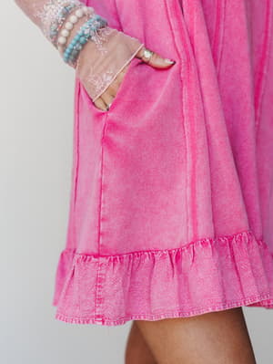 A close-up detail shot of a washed fuschia dress with a ruffled hem and side pocket, styled with a sheer lace sleeve peeking in and a stack of beaded bracelets — the kind of outfit that feels playful and lived-in all at once.