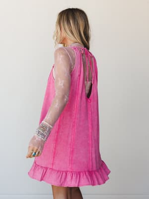 A back-view shot of a washed fuchsia mini dress with an open back and ruffle hem, layered over a sheer floral lace long-sleeve top — the kind of outfit that looks effortless but definitely took thought.