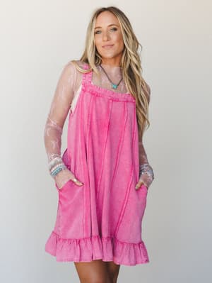 A mineral-washed fuschia overall-style mini dress with a ruffled hem is layered over a sheer floral lace long-sleeve top — the kind of outfit that looks effortless but is clearly very much on purpose.