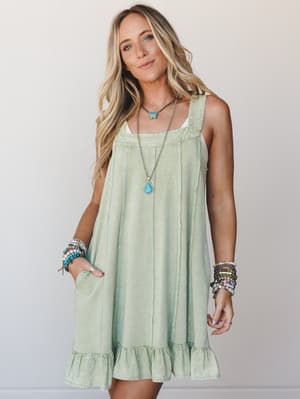 A flowy sage mini dress with a relaxed A-line silhouette and ruffle hem, styled with stacked boho bracelets and a turquoise pendant necklace — the kind of outfit that feels like a deep breath.