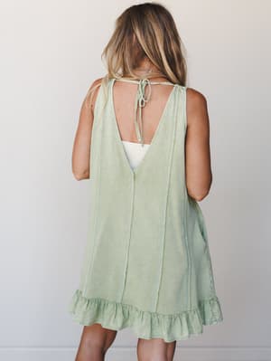 A breezy mini dress in washed sage green shot from the back, showing off its open V-back with tie closure and ruffle hem — the kind of dress that looks like it was made for a slow summer afternoon.