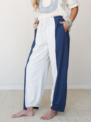 A relaxed, mid-shot of a model wearing color-blocked barrel pants in heathered grey and navy with a drawstring waist, styled with a cream embroidered top and stacked beaded bracelets — easy, lived-in energy with just enough intention.