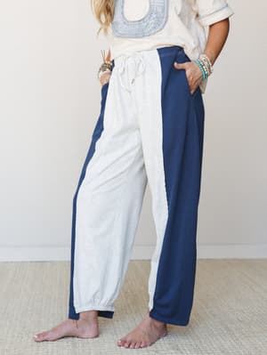 A relaxed, mid-shot of a model wearing color-blocked barrel pants in heathered grey and navy with a drawstring waist, styled with a cream embroidered top and stacked beaded bracelets — easy, lived-in energy with just enough intention.