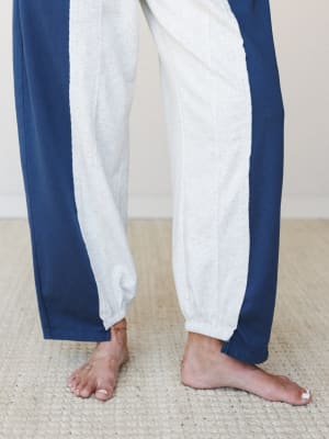 A close-up of relaxed barrel-leg sweatpants in heathered grey with navy side panels, worn barefoot on a woven rug — the kind of pants that make staying in feel like a whole vibe.