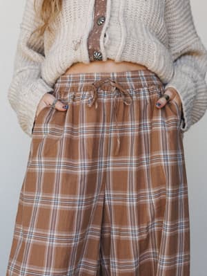 A close-up mid-shot of wide-leg plaid pants in a warm latte brown, styled with a cropped cream knit cardigan and a floral button placket underneath — the kind of outfit that feels like a slow Saturday morning made wearable.