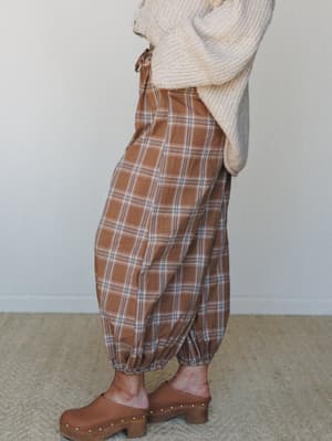 A side-profile shot of relaxed plaid balloon pants in warm latte brown, styled with a chunky cream knit sweater and wood-soled clogs — the kind of autumn outfit you'd wear to the farmers market and never want to take off.
