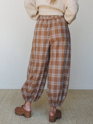 Shot from behind, a model wears voluminous plaid balloon pants in warm caramel tones paired with a chunky cream knit sweater and wood-studded clogs — the whole look reads like a cozy autumn Saturday with somewhere worth going.