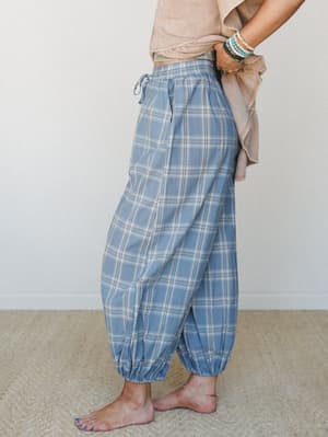 A side-profile shot of relaxed plaid harem pants in steel blue, styled barefoot with a cropped sand-washed tee and stacked turquoise bracelets — the kind of outfit that makes a slow morning feel intentional.
