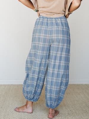 A relaxed back-facing shot of flowy plaid harem pants in washed steel blue, paired with a casual sand tee and stacked bracelets — the kind of pants that make staying home feel like a whole personality.