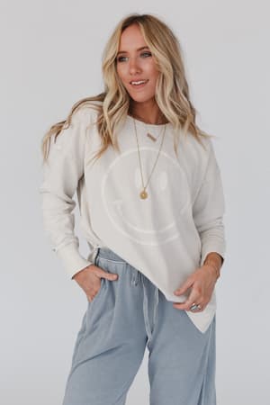 A woman with long blonde hair wears the Catch My Smile Pullover Top in khaki and light blue pants, accessorizing her relaxed fit top with layered necklaces and rings as she poses with one hand in her pocket.