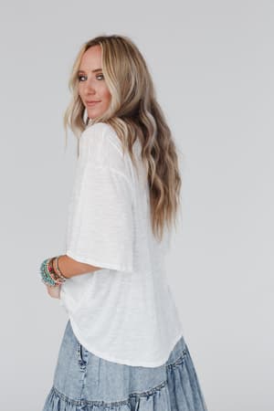 A woman with long, wavy blonde hair wears The Nest Your Favorite Slub Tee Shirt in Natural and a light blue denim skirt, standing sideways and looking over her shoulder, wearing colorful beaded bracelets against a plain light background.