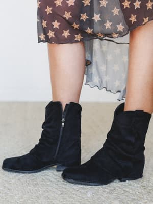 A close-up shot of black suede slouchy ankle booties styled with a sheer star-print midi skirt, shot against a neutral interior backdrop — the kind of effortless pairing that makes you want to get dressed.