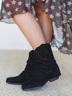 A close-up of black suede slouchy ankle boots styled with a sheer star-print midi skirt, shot from the knees down — the kind of pairing that makes a simple boot feel like a whole personality.