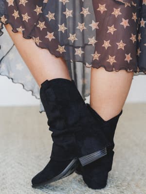 A close-up of black suede slouchy booties with a stacked heel paired with a sheer dark star-print midi skirt, shot with soft natural light — the kind of outfit that feels effortlessly put-together without trying too hard.