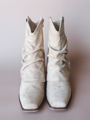 A pair of sand-toned suede slouch booties shot straight-on against a soft lavender-gray studio backdrop — the kind of boots that look equally at home tucked into jeans or peeking out from a flowy midi skirt.