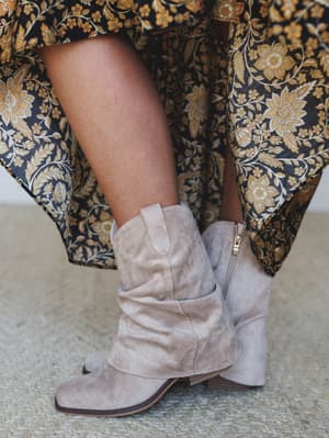 A close-up detail shot of slouchy suede ankle booties in a warm sand tone, paired with a flowy floral-print maxi skirt in navy and gold — the kind of pairing that makes a simple outfit feel intentional.