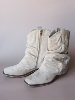 A pair of sand suede slouchy ankle boots with a lived-in, effortlessly rumpled shaft, square toe, and gold side zipper — shot clean against a soft mauve-gray studio backdrop.