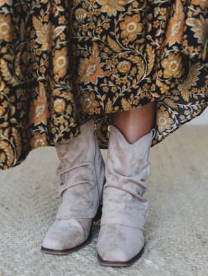 A close-up of slouchy sand suede cowboy-style booties peeking out beneath a sweeping floral maxi skirt in navy and gold — the kind of pairing that looks effortless but was absolutely intentional.