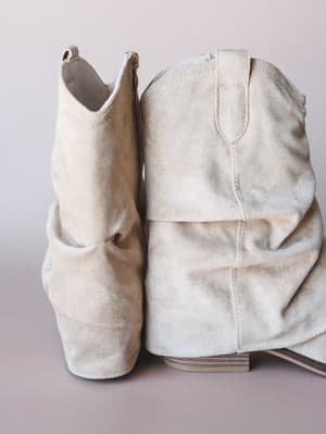 A pair of sand-toned suede slouchy booties shot up close against a soft lavender-grey backdrop — the kind of boots that feel like they were made to be broken in and worn everywhere.