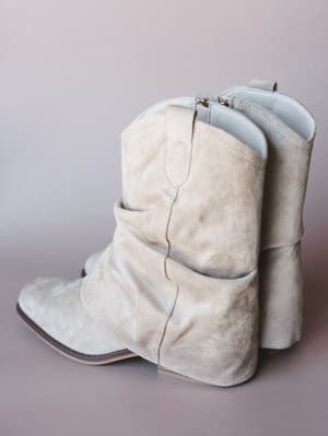 A pair of sand-colored suede slouchy western booties photographed against a soft mauve-gray backdrop — the kind of boots that go with everything and make even a Tuesday feel intentional.