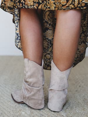 A close-up detail shot from behind, pairing sand suede slouchy cowboy-style booties with a flowing floral-print skirt in navy and gold — the kind of combo that looks effortless but is very much intentional.