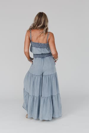 A woman with long blonde hair stands facing away, wearing the Harlow Wide Leg Chambray Jumpsuit - Blue with spaghetti straps and bracelets on her wrists, set against a plain light background.