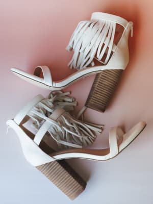 A pair of cream fringe block-heel sandals styled in an overlapping flat lay against a soft blush-to-white gradient background — equal parts boho and going-out energy.